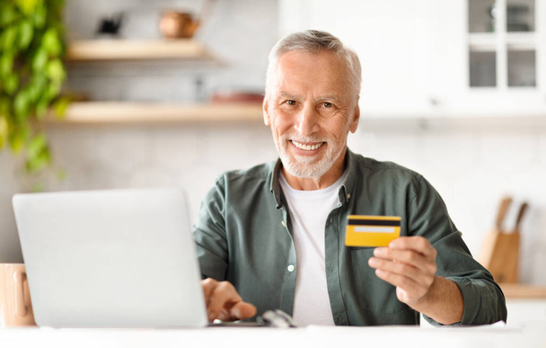 E-Ticaret Konsepti. Gülümseyen kıdemli adam kredi kartını tutuyor ve evde dizüstü bilgisayar kullanıyor, mutlu yaşlı adam çevrimiçi ödeme veya internet alışverişi yapıyor, web bankacılığının tadını çıkarıyor, kopyalama alanı - Fotoğraf, Görsel