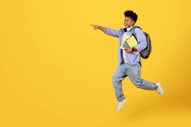 Energetic young black male student with headphones and backpack leaping in mid-air, pointing forward at free space with big smile, against striking yellow backdrop - Foto, Imagem