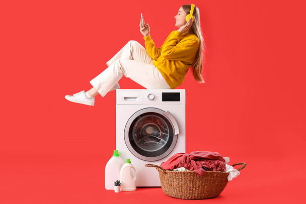 Hübsche junge Frau sitzt auf Waschmaschine und hört Musik vor rotem Hintergrund - Foto, Bild