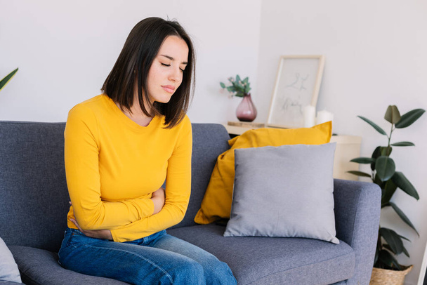 Young woman suffering stomach ache or abdominal pain while sitting on couch at home. Menstruation or pregnancy health care effects - Photo, Image