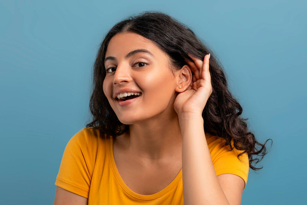 Взволнованная счастливая молодая кудрявая индианка в жёлтой футболке, держащая за руку рядом с ушным подслушиванием, изолированная на синем фоне студии. Жест и эмоции - Фото, изображение