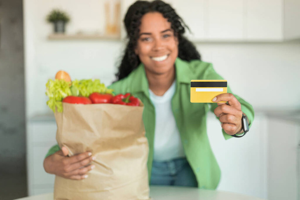 Mujer negro alegre sostiene tarjeta de crédito y bolsa de papel con productos de alimentación en la cocina moderna, celebra la conveniencia de compras en línea y la alegría de una nutrición saludable. Enfoque selectivo - Foto, Imagen