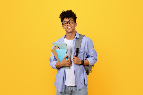 glimlachende jonge zwarte mannelijke student in bril dragen notebooks en rugzak staat tegen gele achtergrond en glimlachen op camera - Foto, afbeelding