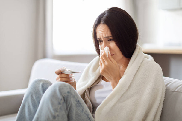 Hasta genç bir kadın burnunu sümkürürken ve evde termometreye bakarken kadın sıcaklığı kontrol ediyor ve oturma odasında battaniyeye sarılmış bir şekilde peçeteye hapşırıyor. - Fotoğraf, Görsel