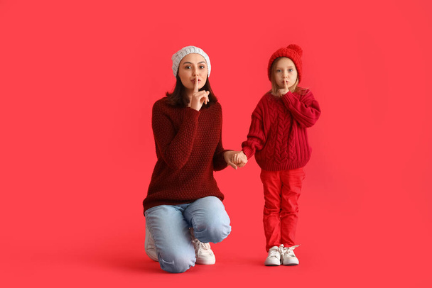 Young mother and her little daughter showing silence gesture on red background - Foto, imagen