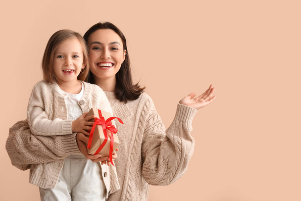 Young mother and her little daughter with Christmas gift showing something on beige background - Photo, Image