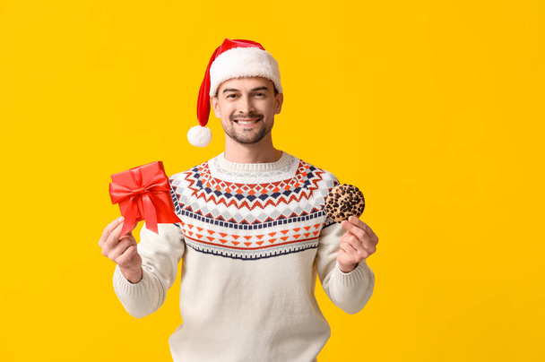 Handsome man in Santa hat with gift box and tasty cookie on yellow background - Photo, Image
