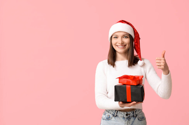 Beautiful young woman in Santa hat with gift boxes showing thumb-up on pink background - Photo, Image