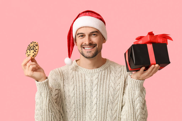 Schöner Mann mit Weihnachtsmütze mit Geschenkschachteln und leckerem Plätzchen auf rosa Hintergrund - Foto, Bild