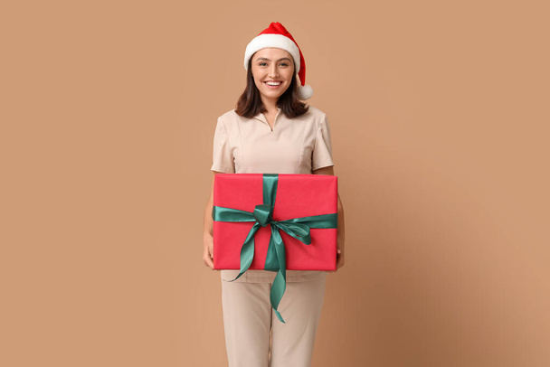 Beautiful female massage therapist in Santa hat with gift box on brown background - Photo, Image