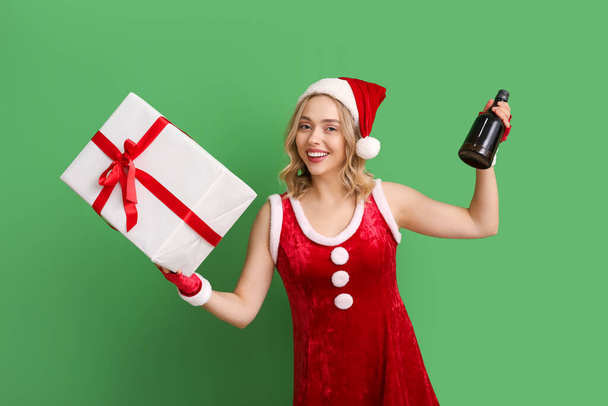 Belle jeune femme habillée en Père Noël avec cadeau de Noël et bouteille de champagne sur fond vert - Photo, image