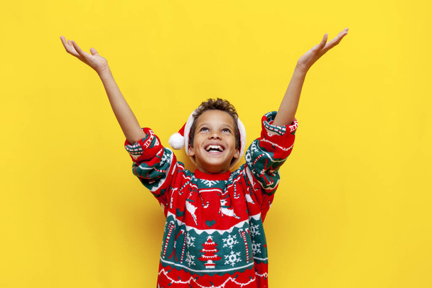 menino americano africano alegre em roupas de Natal mantém as mãos vazias e olha para cima no fundo isolado amarelo, criança de 9 anos em sonhos de santa hat e captura espaço de cópia - Foto, Imagem