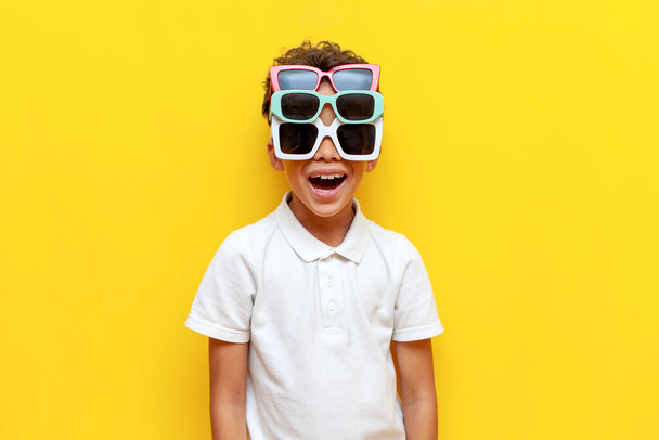 schockierter afrikanisch-amerikanischer Junge mit drei Sonnenbrillen, die Überraschung auf gelbem, isoliertem Hintergrund zeigen, lustiger Teenager mit verschiedenen Brillen, der Erstaunen zeigt - Foto, Bild