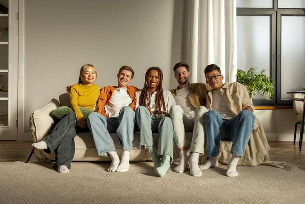 πολυφυλετική ομάδα νεαρών φίλων που κάθονται στον καναπέ στο σπίτι αγκαλιάζοντας και κοιτάζοντας την κάμερα, ομάδα μαθητών που βλέπουν τηλεόραση μαζί στο δωμάτιο και χαμογελώντας - Φωτογραφία, εικόνα