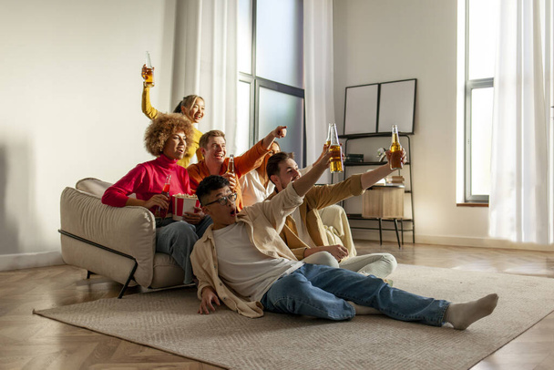 multiracial group of young friends sitting on the couch at home drinking beer and watching TV and celebrating good luck, students drinking alcohol rejoicing in victory and cheering and screaming - Foto, Bild