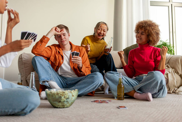 πολυφυλετική ομάδα νεαρών φίλων που παίζουν χαρτιά πίνοντας μπύρα και διασκεδάζοντας στο σπίτι, μαθητές που κάθονται στο πάτωμα και παίζουν τυχερά παιχνίδια και πίνουν αλκοόλ μαζί στο δωμάτιο - Φωτογραφία, εικόνα
