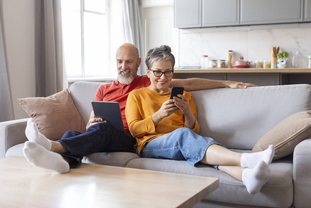 Vieux couple souriant reposant sur un canapé avec tablette numérique et téléphone portable, heureux homme et femme âgés utilisant les technologies modernes à la maison, se détendre avec des gadgets, profiter des loisirs de retraite - Photo, image