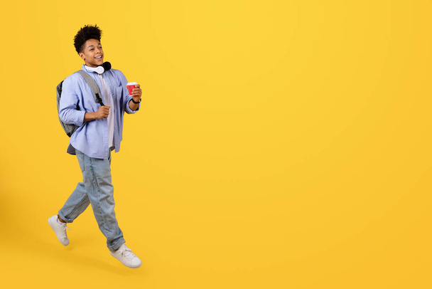 Wesoły czarny student w luźnym ubraniu, ze słuchawkami i plecakiem, trzymając filiżankę kawy podczas spaceru i patrząc w bok, ustawiony na jaskrawym żółtym tle - Zdjęcie, obraz