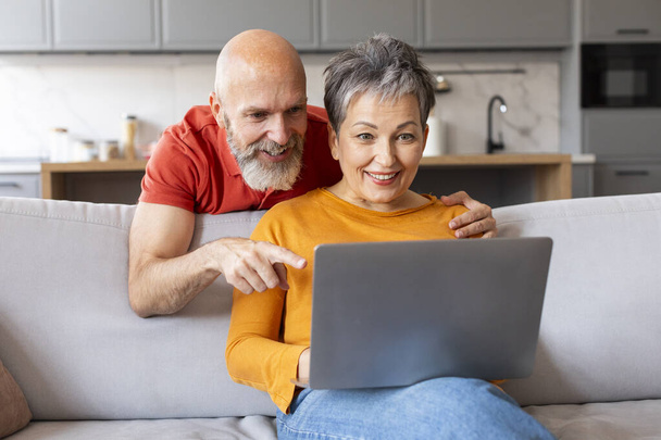 Oturma odasındaki koltukta dinlenirken dizüstü bilgisayarlı mutlu yaşlı çift internetten alışveriş yaparken ya da tatil yaparken gülümsüyor. - Fotoğraf, Görsel