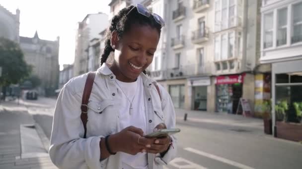 Schöne junge schwarze Frau, die mit Handy in der Hand die Straße hinuntergeht, sie schaut lächelnd und lustig auf den Telefonbildschirm, plaudert und sendet Sprachaudios durch die Stadt. - Filmmaterial, Video