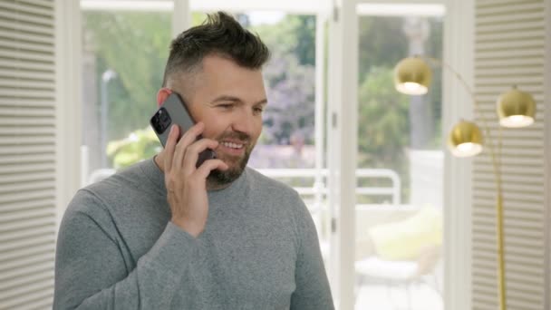 Positiivinen mies soittaa matkapuhelimeen seisoo modernissa kaupunkiasunnossa. Komea mies nauraa älypuhelimella sisätiloissa. Hymyilevä kolmekymppinen mies rennossa asussa. Älypuhelintekniikka - Materiaali, video