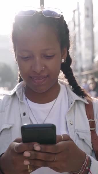Κάθετη βίντεο από μια όμορφη νεαρή μαύρη γυναίκα στέκεται στο δρόμο με το κινητό στα χέρια της, γυρίζει κοιτάζει κάμερα και χαμογελά και αστείο με ευτυχισμένη έκφραση. Latina κορίτσι σε εξωτερικούς χώρους στην πόλη. - Πλάνα, βίντεο