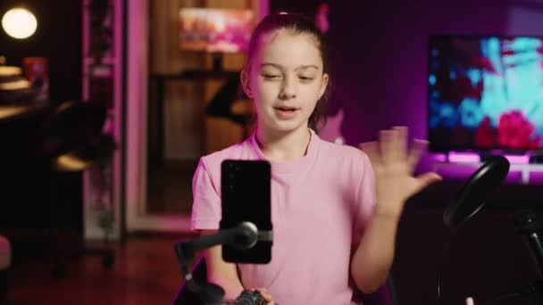 Kid films with smartphone attached to selfie stick in pink neon lit living room used as professional studio. Young media star captures footage with cellphone camera, discussing about fun day at school - Footage, Video