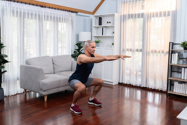 Actief en fit senior man opwarmen en stretching voor thuis oefening routine in de woonkamer. Gezond fitness lifestyle concept na pensionering voor gepensioneerden. Klok - Foto, afbeelding