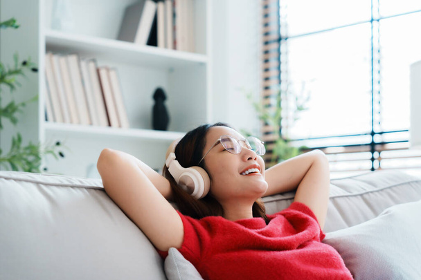 Mujer asiática joven y relajada disfrutando del descanso en un cómodo sofá, tranquila chica atractiva relajándose en el sofá, respirando aire fresco con los ojos cerrados, usando anteojos y auriculares, meditando en casa - Foto, Imagen