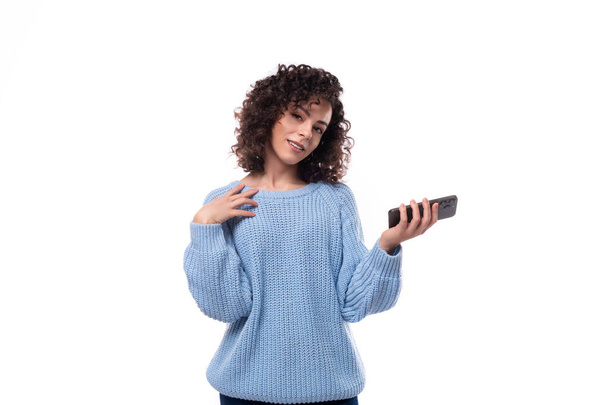 pretty young curly brunette woman dressed in a blue sweater holding a smartphone. - Photo, Image
