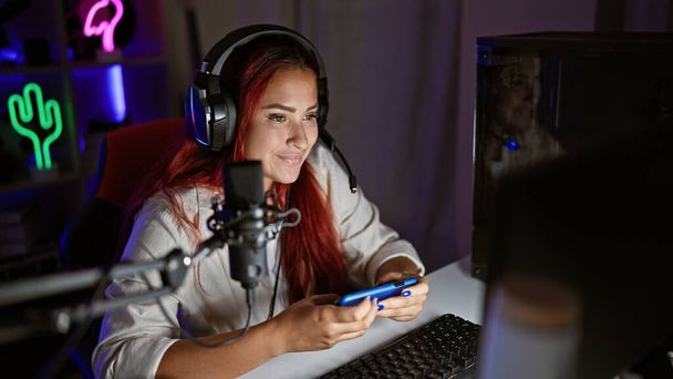 活気に満ちた若い赤毛の女性ストリーマーは,暗いゲームルームでスマートフォンで仮想ゲームをうまく再生 - 写真・画像