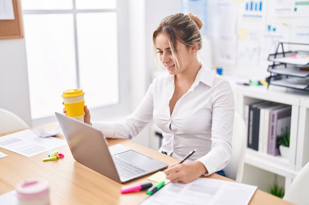 Young blonde woman business worker writing on reminder paper holding coffee at office - Photo, Image
