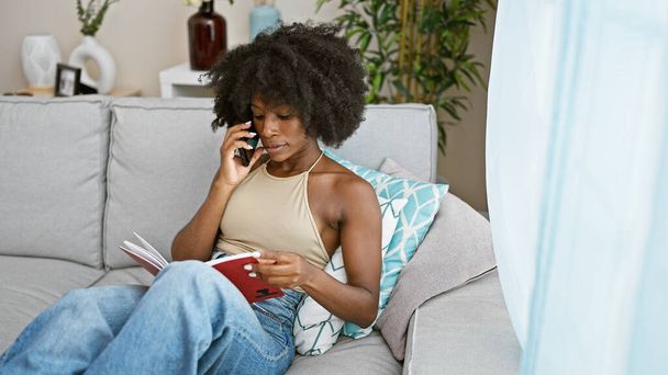 African american woman talking on smartphone reading book at home - Photo, Image