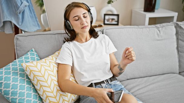 Молодая женщина слушает музыку, сидя на диване, танцуя дома - Фото, изображение