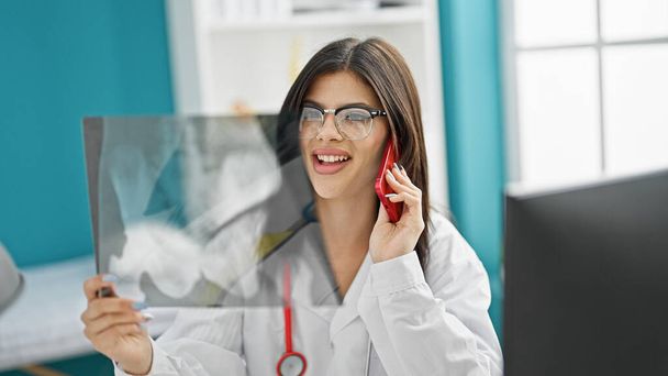Νεαρή καυκάσια γυναίκα γιατρός μιλάει στο smartphone κοιτάζοντας ακτινογραφία στην κλινική - Φωτογραφία, εικόνα