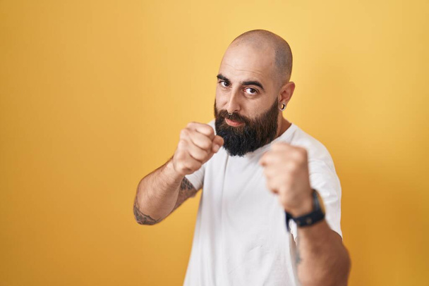Jeune homme hispanique avec barbe et tatouages debout sur fond jaune prêt à se battre avec geste de défense poing, visage en colère et bouleversé, peur du problème  - Photo, image