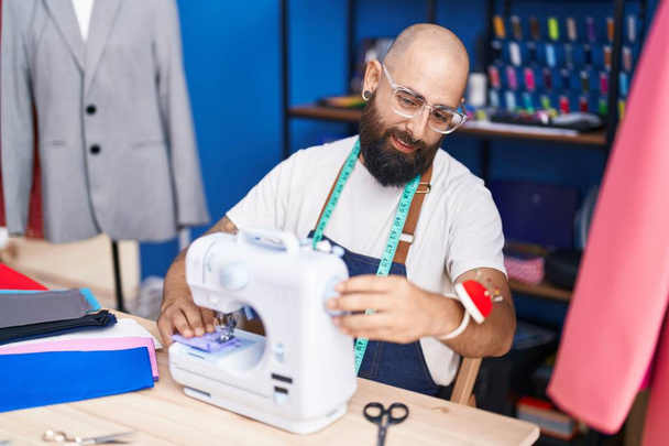 Jovem careca alfaiate sorrindo confiante usando máquina de costura na fábrica de roupas - Foto, Imagem