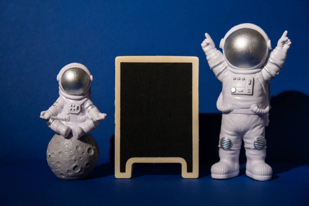 Plastic toy astronaut with blackboard template mock up for your text on colorful blue background Copy space. Concept of out of earth travel, private spaceman commercial flights. Space missions and Sustainability - Photo, image