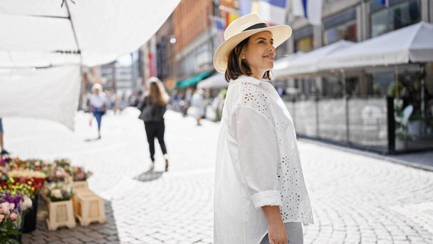 Beautiful young hispanic woman smiling confident looking to the side wearing summer hat at city market - Photo, Image