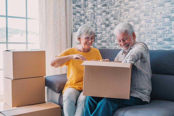 Relaxed mature married couple in love resting on couch among paper cardboard boxes, taking break, pause, hugging, talking, enjoying being in new home. Real estate concept - Photo, Image
