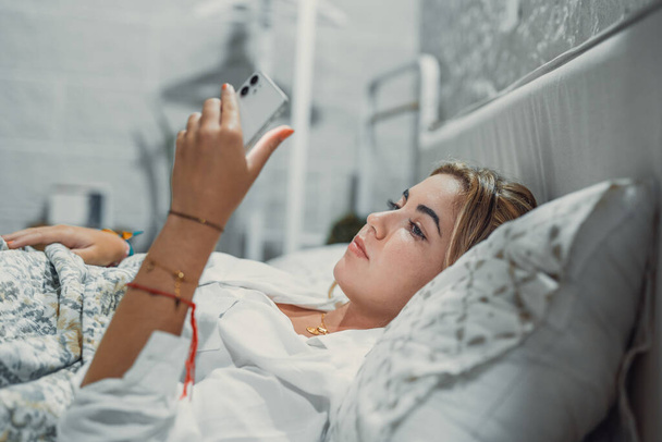 Gadget addiction. Calm bored young woman or teenage girl lying at cozy bed holding cellphone in hands looking at screen chatting checking social network account before fall asleep or after waking up - Photo, Image