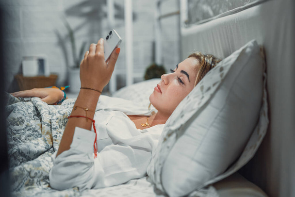 Uzależnienie od gadżetów. Spokojny znudzony młoda kobieta lub nastolatka leżąca w przytulnym łóżku trzymając telefon komórkowy w rękach patrząc na ekranie czatu sprawdzając konto w sieci społecznościowej przed zasnąć lub po przebudzeniu - Zdjęcie, obraz