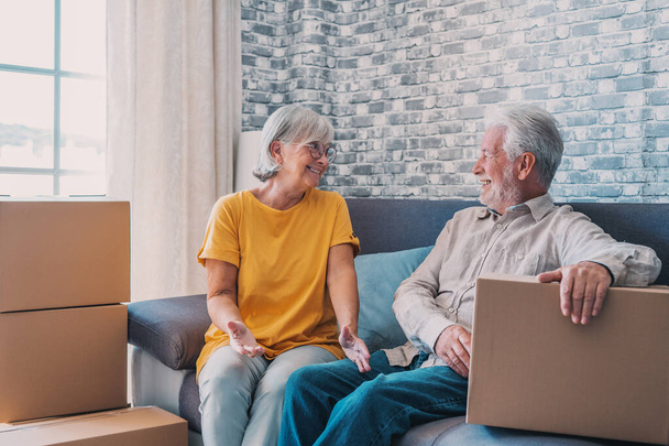 Relaxed mature married couple in love resting on couch among paper cardboard boxes, taking break, pause, hugging, talking, enjoying being in new home. Real estate concept - Photo, Image