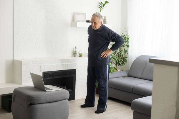 Sport in mature age. Happy middle-aged man doing stretching exercises in front of laptop at home, watching online tutorials, in living room interior, free space. - Foto, imagen