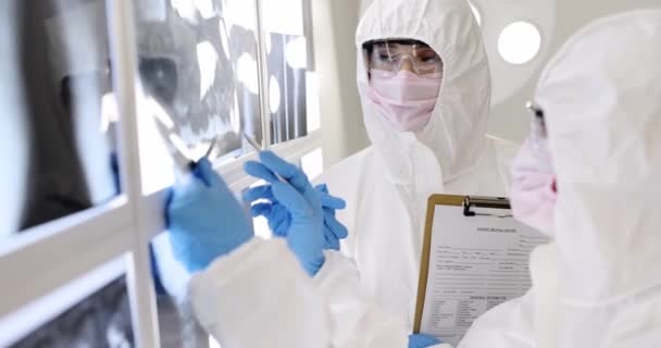 Zwei Ärzte in Schutzanzügen, Masken und Schutzbrillen senden Röntgenstrahlen von Patienten aus. Coronavirus-Infektion und Lungenentzündung - Filmmaterial, Video
