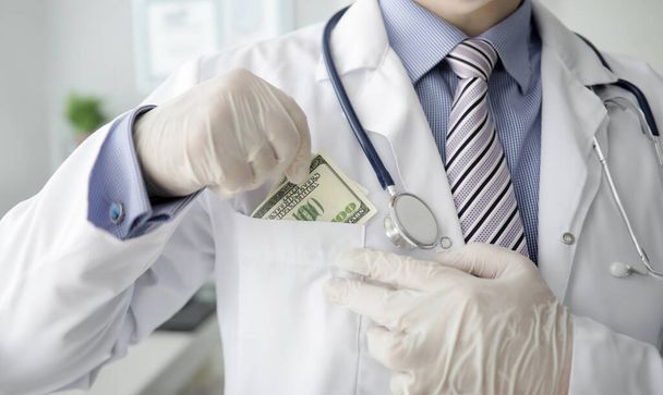 La mano masculina del doctor con guantes blancos mete sobornos de dólares en el bolsillo del uniforme. Concepto de seguro de vida médico
 - Foto, imagen