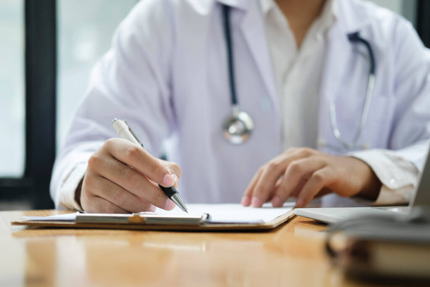 Hands of general practitioner filling paper medical records. Doctor in white coat doing paperwork at workplace with laptop, writing notes, preparing documents, reports, prescription. - Photo, Image