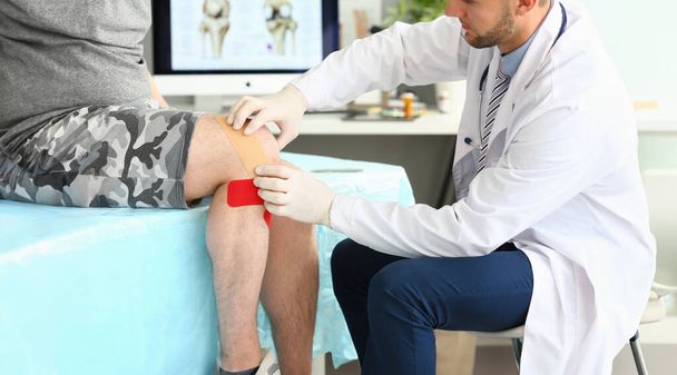 Close-up of smart physician applying recovery tape on mans injured leg. Hurt knee bound with red health care and protective, sticky red bandages. Medical treatment concept - Photo, Image