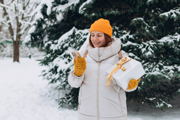 Πορτρέτο του χαμογελαστού κομψή γυναίκα 30 με το παρόν έξω στο πάρκο της πόλης το χειμώνα. Νεαρές γυναίκες με τηλέφωνο κρατώντας χριστουγεννιάτικο δώρο σε εξωτερικούς χώρους - Φωτογραφία, εικόνα
