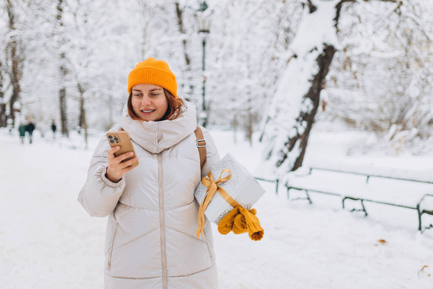 Portret uśmiechniętej kobiety po trzydziestce z obecną zimą w parku miejskim. Młode kobiety z telefonem gospodarstwa Boże Narodzenie obecny na zewnątrz - Zdjęcie, obraz